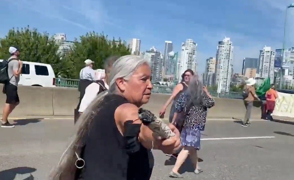 Une femme tient un objet près du micro d’une journaliste de Rebel News sur le pont Cambie, devant la silhouette du centre-ville de Vancouver. Capture du reportage de Rebel News.