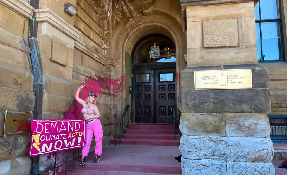 Casey « Ever » Hatherly se tient devant l’entrée du bureau du premier ministre, éclaboussée de peinture rose, avec une pancarte réclamant une action climatique. Photo : Patrick Morrell / CityNews.