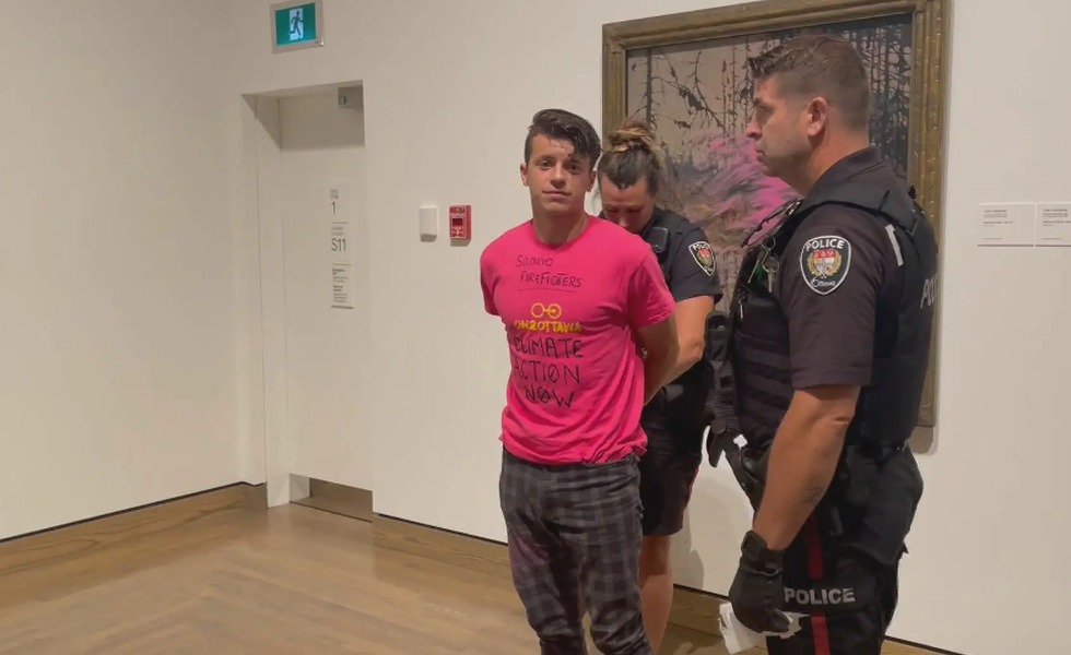 Un jeune homme en chandail rose est escorté par des policiers dans la Galerie nationale après avoir projeté de la peinture sur la vitre protégeant « Northern River ». Photo : On2Ottawa.