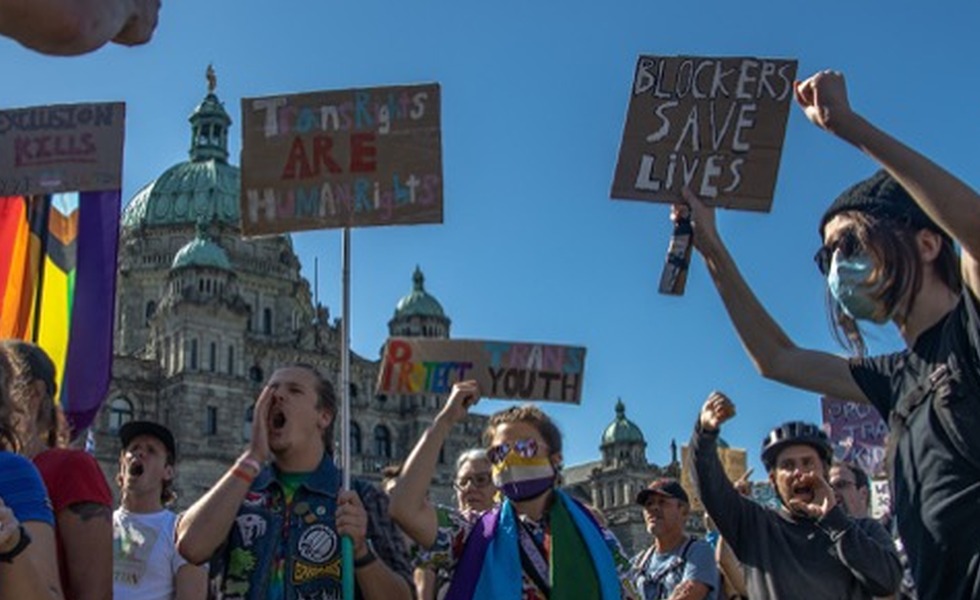 Une foule se rassemble devant l’Assemblée législative de la Colombie-Britannique avec des drapeaux arc-en-ciel et des pancartes, dont « Blockers save lives ». Photo : Robyn Bell / Capital Daily.