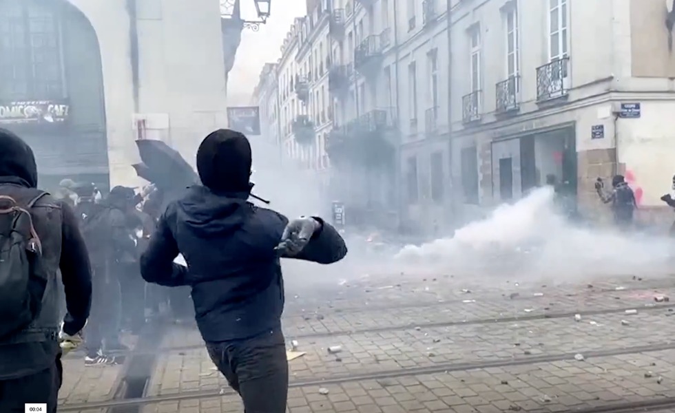 Des manifestants vêtus de noir avancent dans une rue parisienne saturée de gaz lacrymogène; l’un d’eux lance un objet en direction des forces de l’ordre le 23 mars 2023.
