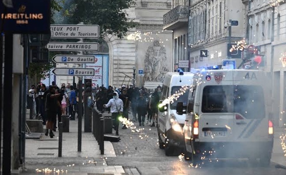 Un fourgon de police avance dans une rue de Marseille jonchée de débris et d’étincelles, tandis que des personnes observent les affrontements depuis les trottoirs.