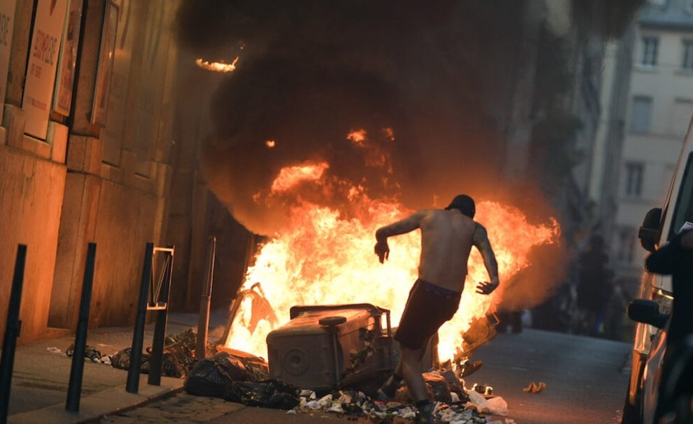 Une personne passe près de conteneurs et de détritus en feu dans une rue de Lyon, où les flammes et la fumée marquent les émeutes de l’été 2023.