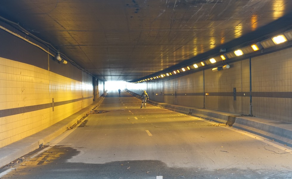 Vue intérieure d’un long tunnel routier carrelé et éclairé, avec une chaussée déserte, quelques débris au sol et deux silhouettes au loin.