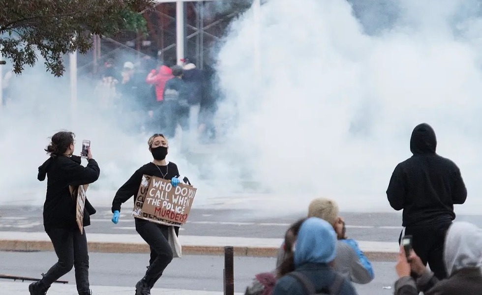 Des manifestants masqués traversent un vaste nuage de gaz lacrymogène au centre-ville de Montréal; l’une d’eux porte une pancarte dénonçant les violences policières. Photo : Graham Hughes / La Presse canadienne, publiée par Global News.