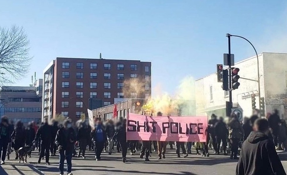 Des manifestants vêtus de noir avancent dans le Mile-Ex derrière une grande bannière rose portant un slogan hostile à la police, au milieu de fumigènes colorés. Photo : soumission anonyme publiée par Montréal Counter-information.