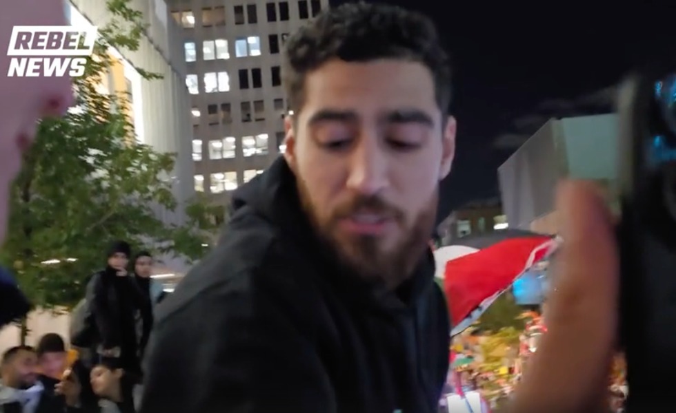 Un homme barbu tend le bras vers la caméra au milieu d’une foule portant des drapeaux palestiniens pendant la mobilisation du 7 octobre 2024. Capture du reportage de Rebel News / YouTube.