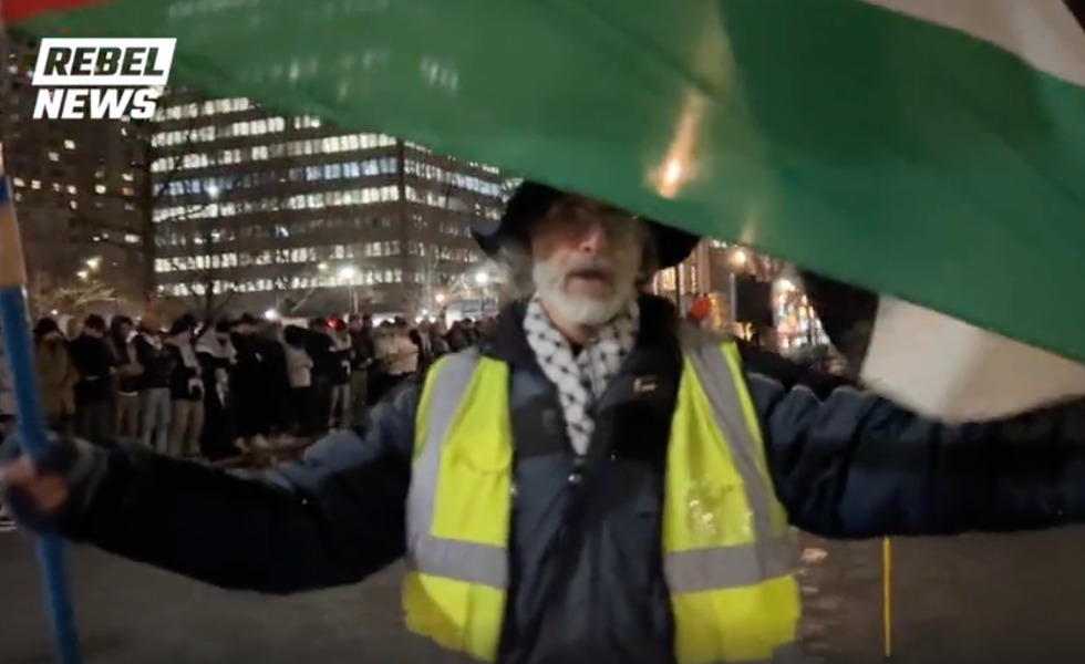 Un homme âgé portant un gilet réfléchissant se tient sous un grand drapeau palestinien au milieu d’un rassemblement nocturne à la Place du Canada. Capture du reportage de Rebel News / YouTube.