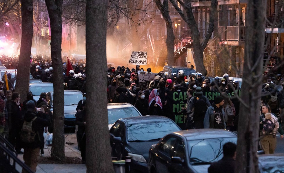 Un important cortège anticapitaliste défile à la tombée du jour entre les arbres, avec des pancartes, des feux rouges et de la fumée en arrière-plan. Photo : Pamela Pagano / CityNews.