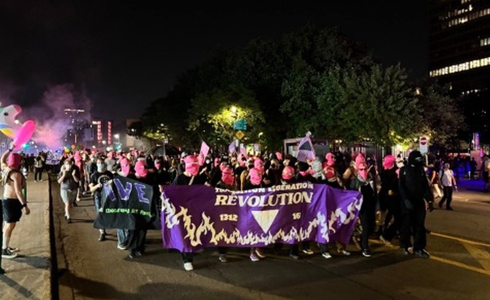 Un cortège nocturne avance derrière une large bannière violette portant le mot « REVOLUTION », sous un éclairage violet. Photo publiée dans la couverture de Rad Pride.