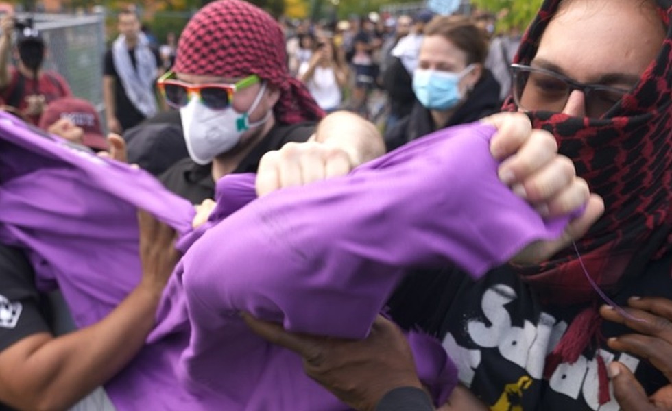 Plusieurs personnes masquées se disputent une étoffe violette au cours d’une bousculade pendant la vigile du 27 septembre 2025 à Longueuil. Capture du reportage de Rebel News / YouTube.