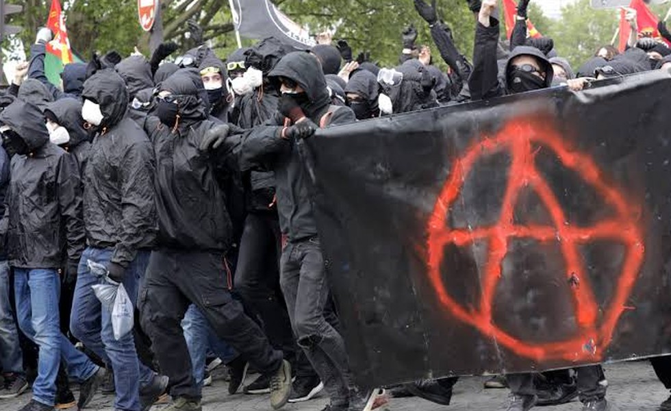 Des manifestants vêtus de noir et le visage couvert tiennent une grande bannière noire marquée d’un symbole anarchiste rouge; des drapeaux sont visibles à l’arrière-plan.