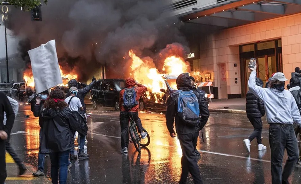 Plusieurs véhicules brûlent dans une rue de Seattle tandis que des manifestants masqués circulent à proximité.