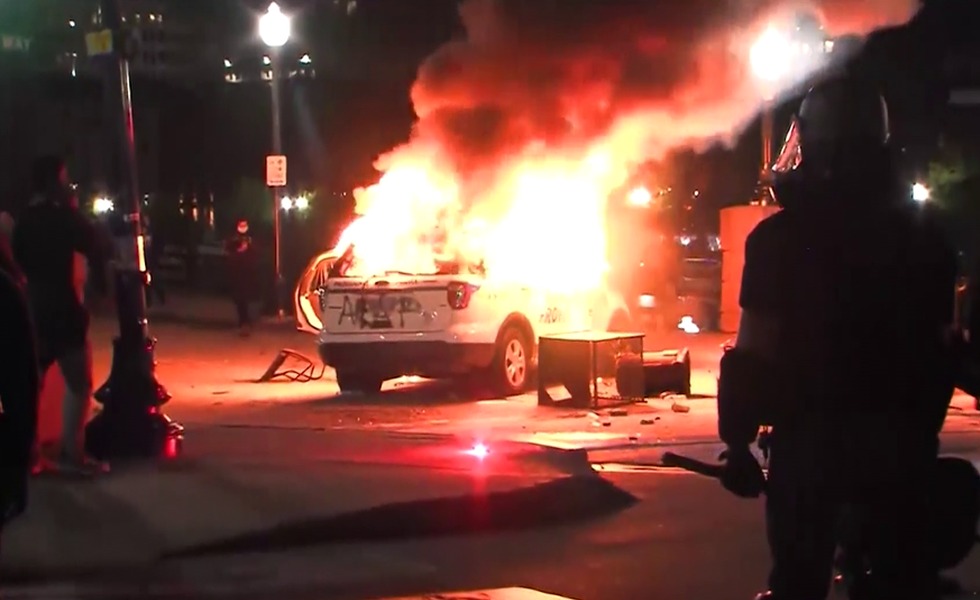 Un véhicule de police est entièrement dévoré par les flammes dans une rue de Providence, sous le regard de personnes rassemblées à proximité.