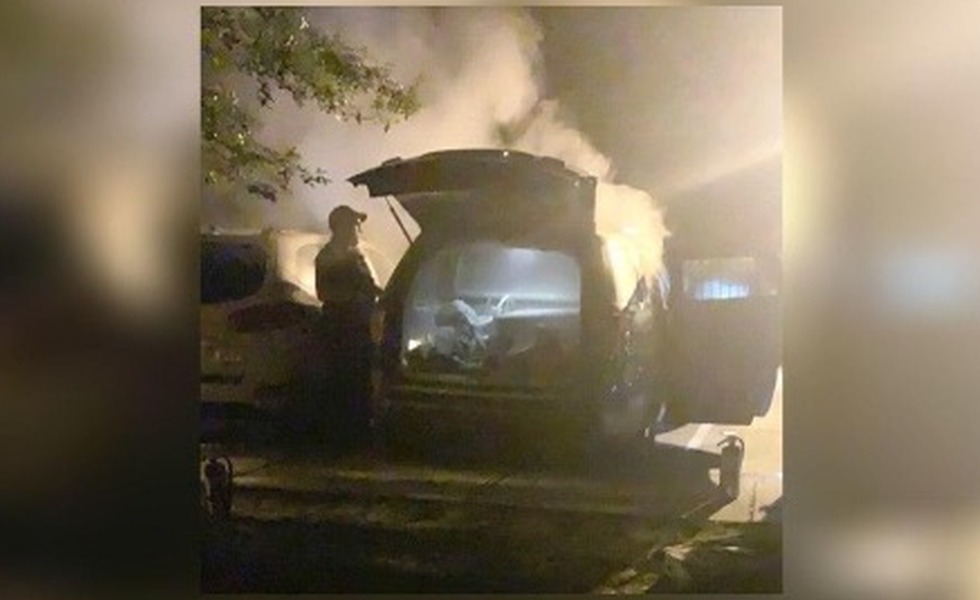 Un véhicule de police endommagé dégage encore de la fumée dans la nuit, tandis qu’une personne se tient près de l’arrière ouvert.