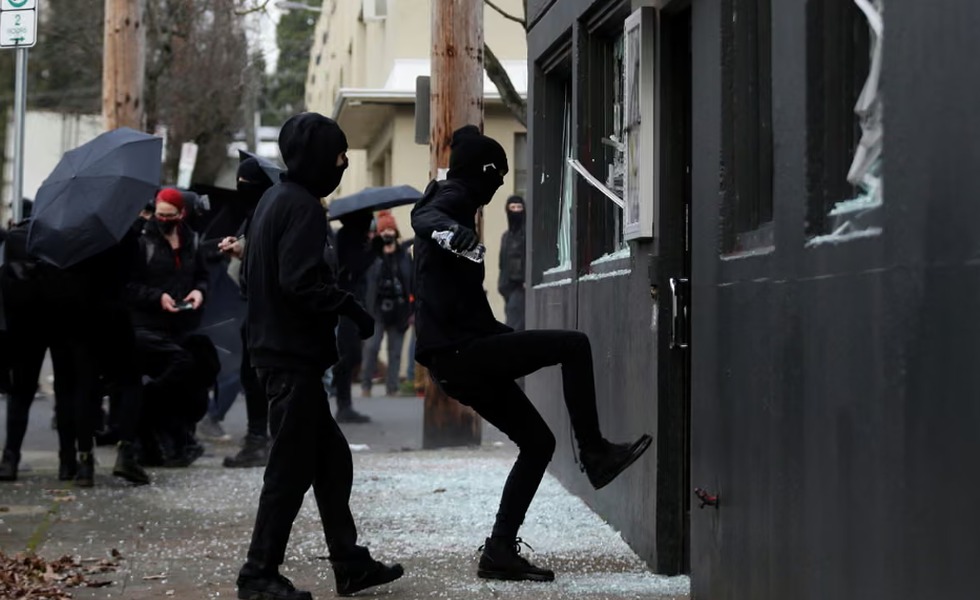 Des personnes vêtues de noir et masquées fracassent les vitrines et la porte d’un bâtiment lors d’une action à Portland.