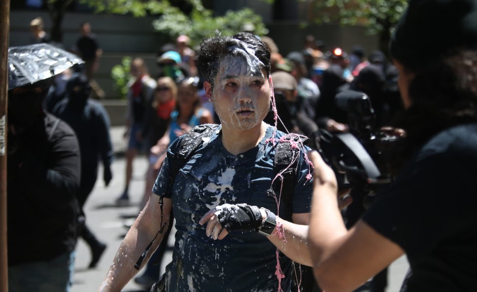 Andy Ngo, le visage et les vêtements couverts d’un liquide blanc, est entouré de personnes vêtues de noir.