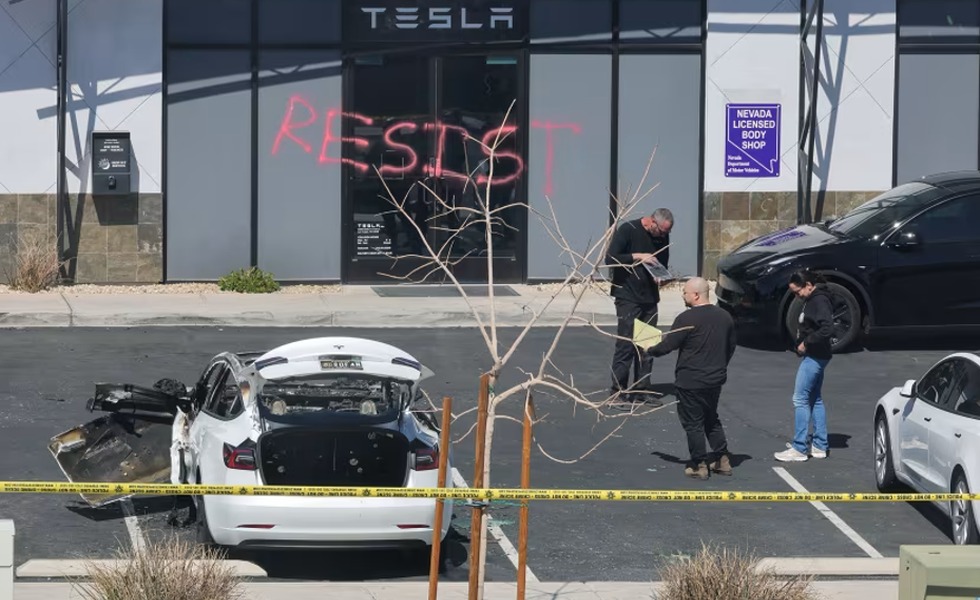 Un centre de service Tesla porte le graffiti « RESIST »; un véhicule blanc endommagé se trouve derrière un ruban de police.